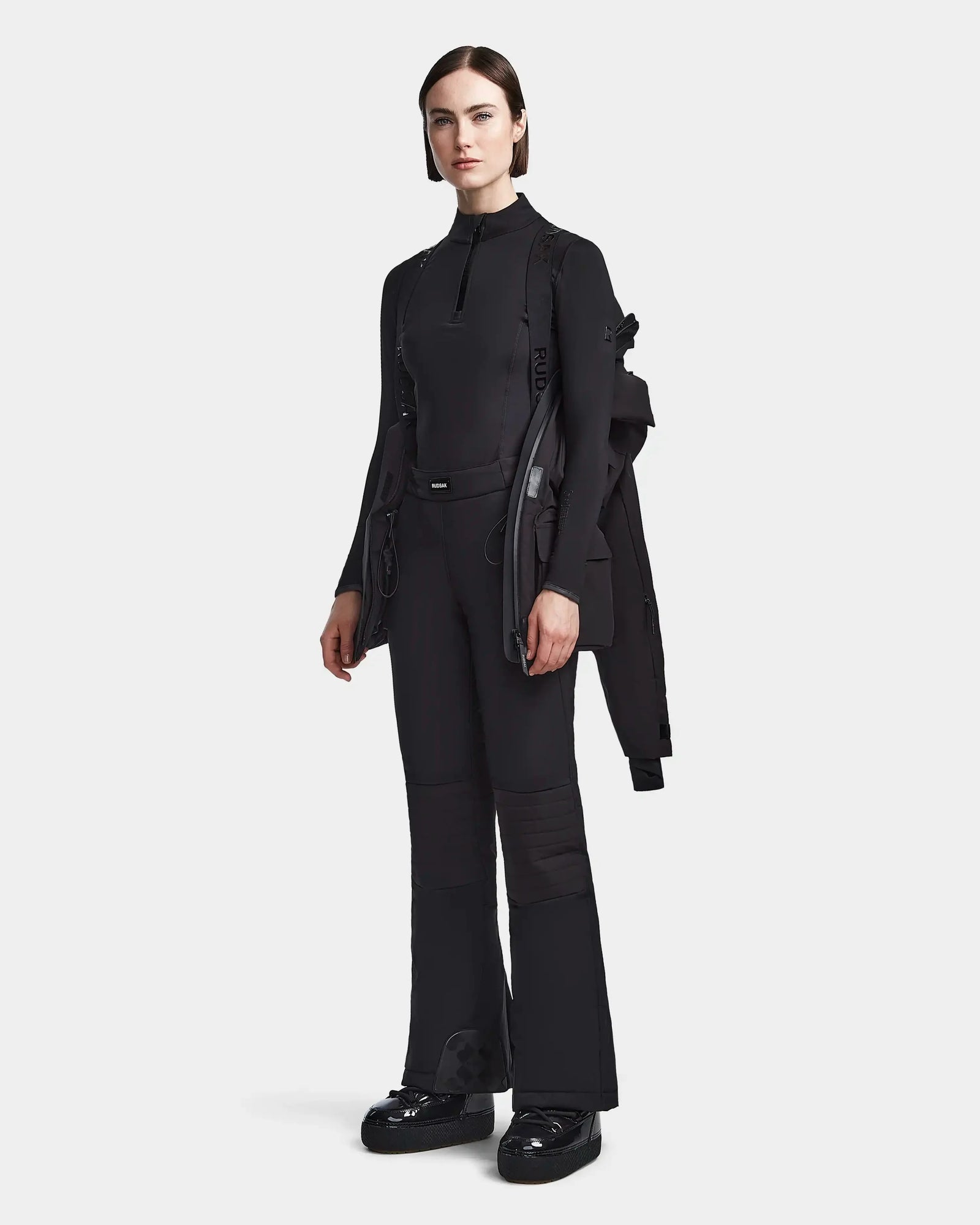 A woman models Rudsaks LUX - 8123618 Black in a sleek, all-black ensemble with flared pants and chunky shoes, holding a womens ski jacket over one shoulder against a light gray background—effortless winter style.
