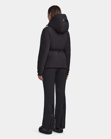 A woman stands facing away in the Rudsak LUX - 8123618 Black womens ski jacket with RECCO technology and matching black pants with zippered bottoms, set against a plain light background, showcasing sleek winter style.
