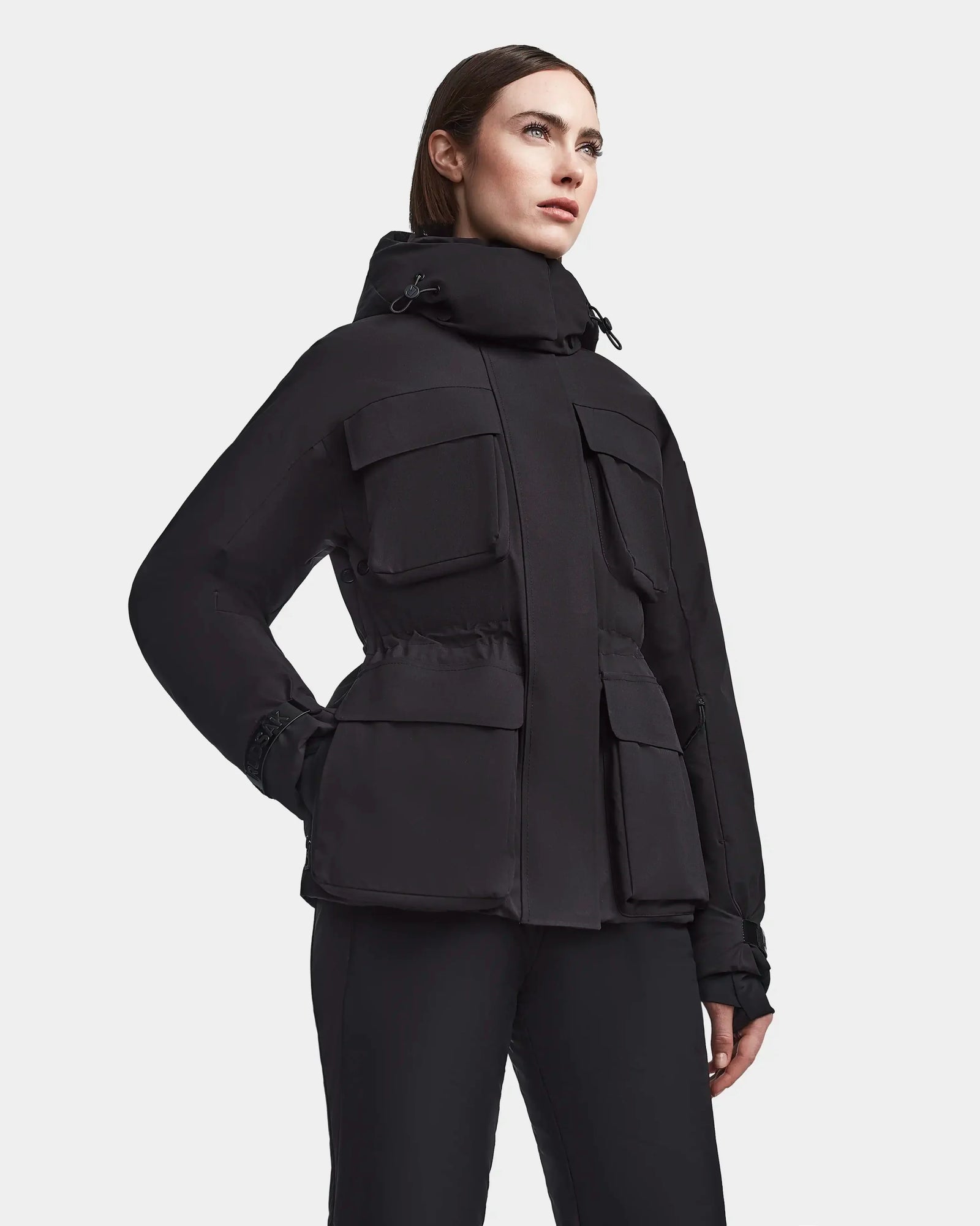 A woman models the Rudsak LUX - 8123618 Black ski jacket, featuring a hood and multiple large front pockets, against a white background—a sleek winter fashion choice.