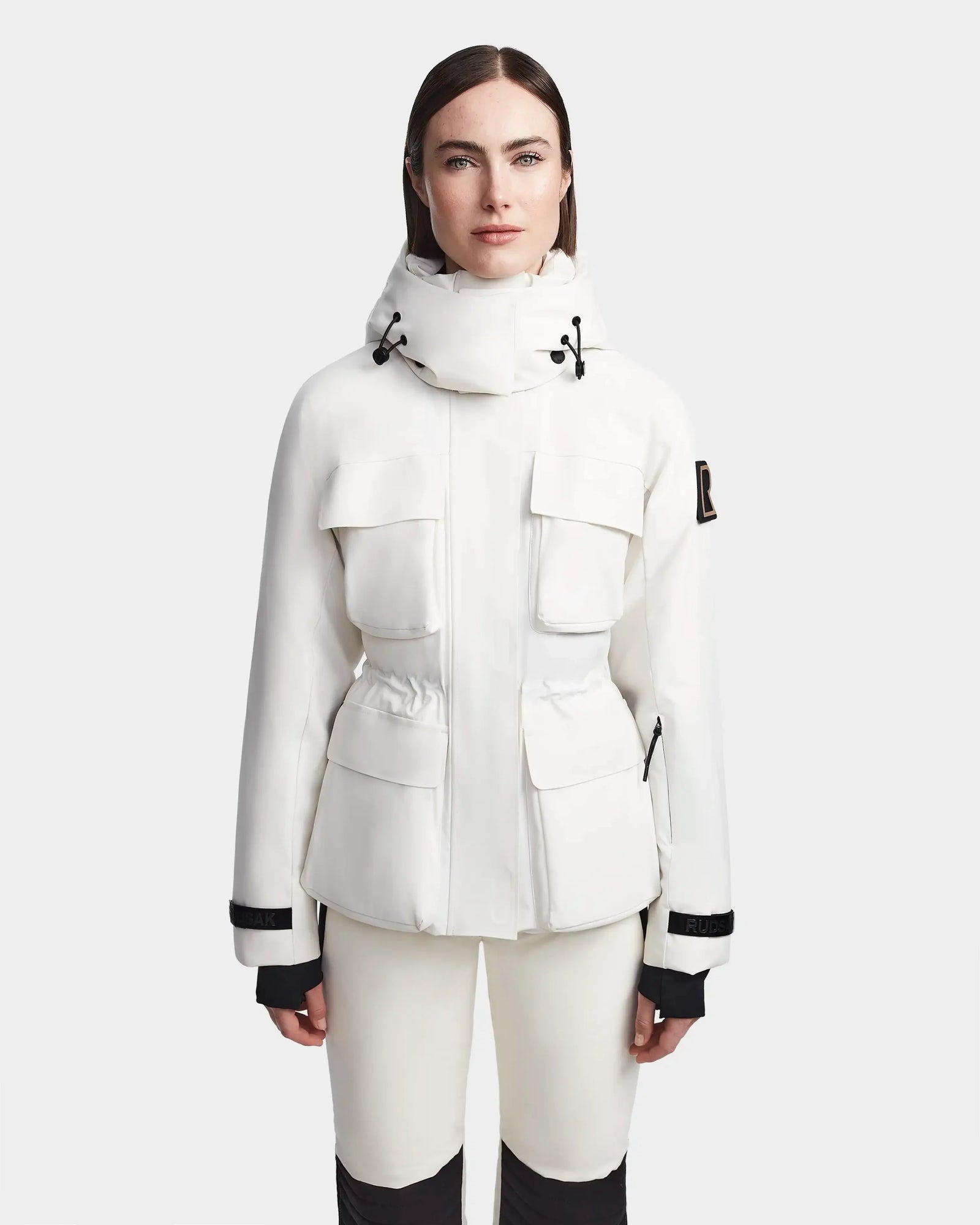 A woman faces forward in a Rudsak LUX - 8123618 Iceland white ski jacket with large front pockets, black trim, and RECCO technology, paired with matching white pants against a plain light background.