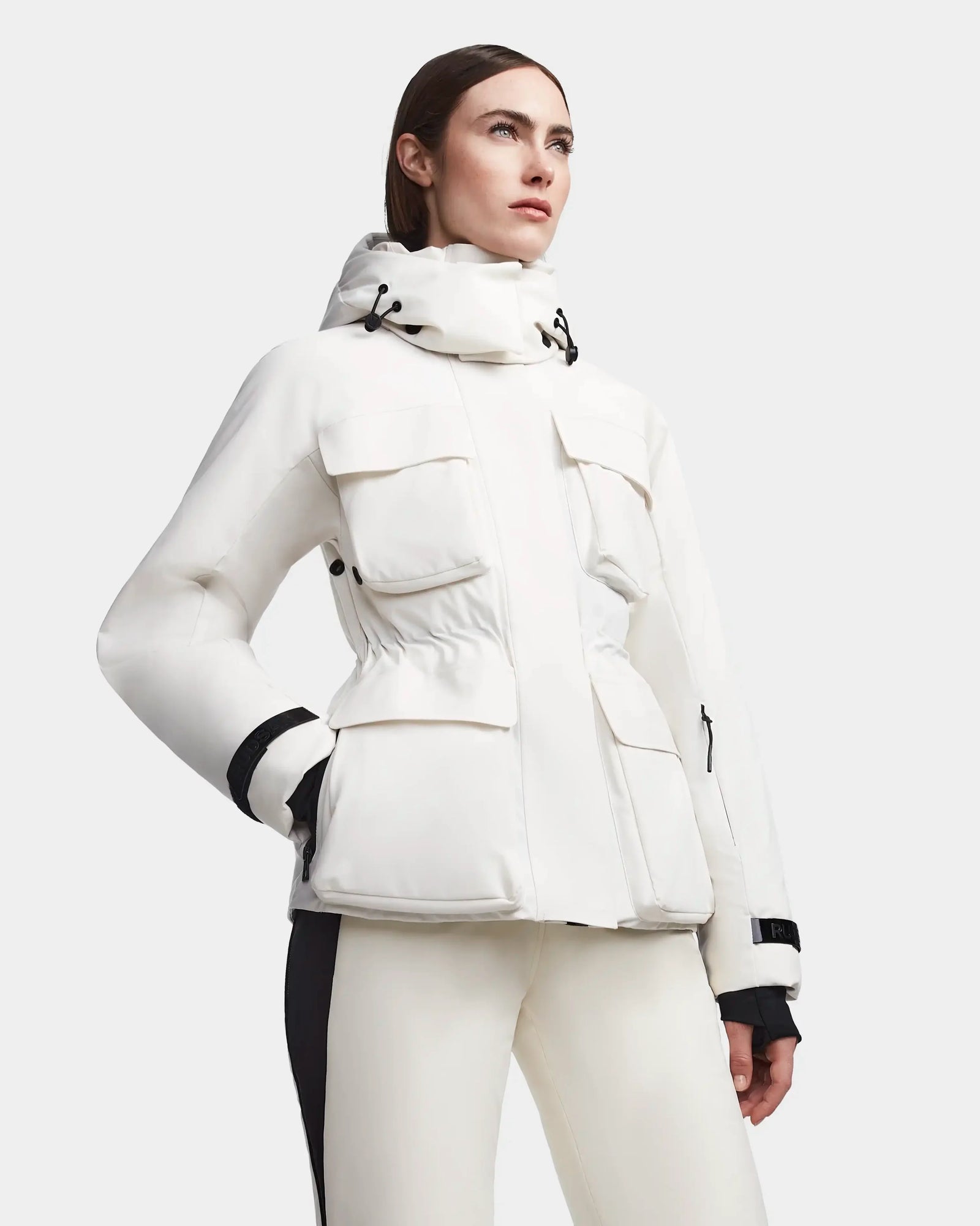 A woman models the Rudsak LUX - 8123618 Iceland white ski jacket with large front pockets and matching pants, posing confidently against a plain light gray background.