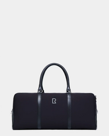 The Rudsak OLIVER - 9323507 Black is a unisex duffel bag with dual handles, vertical leather-like accents, and a central silver geometric logo, offering a luxurious travel look against a plain light background.