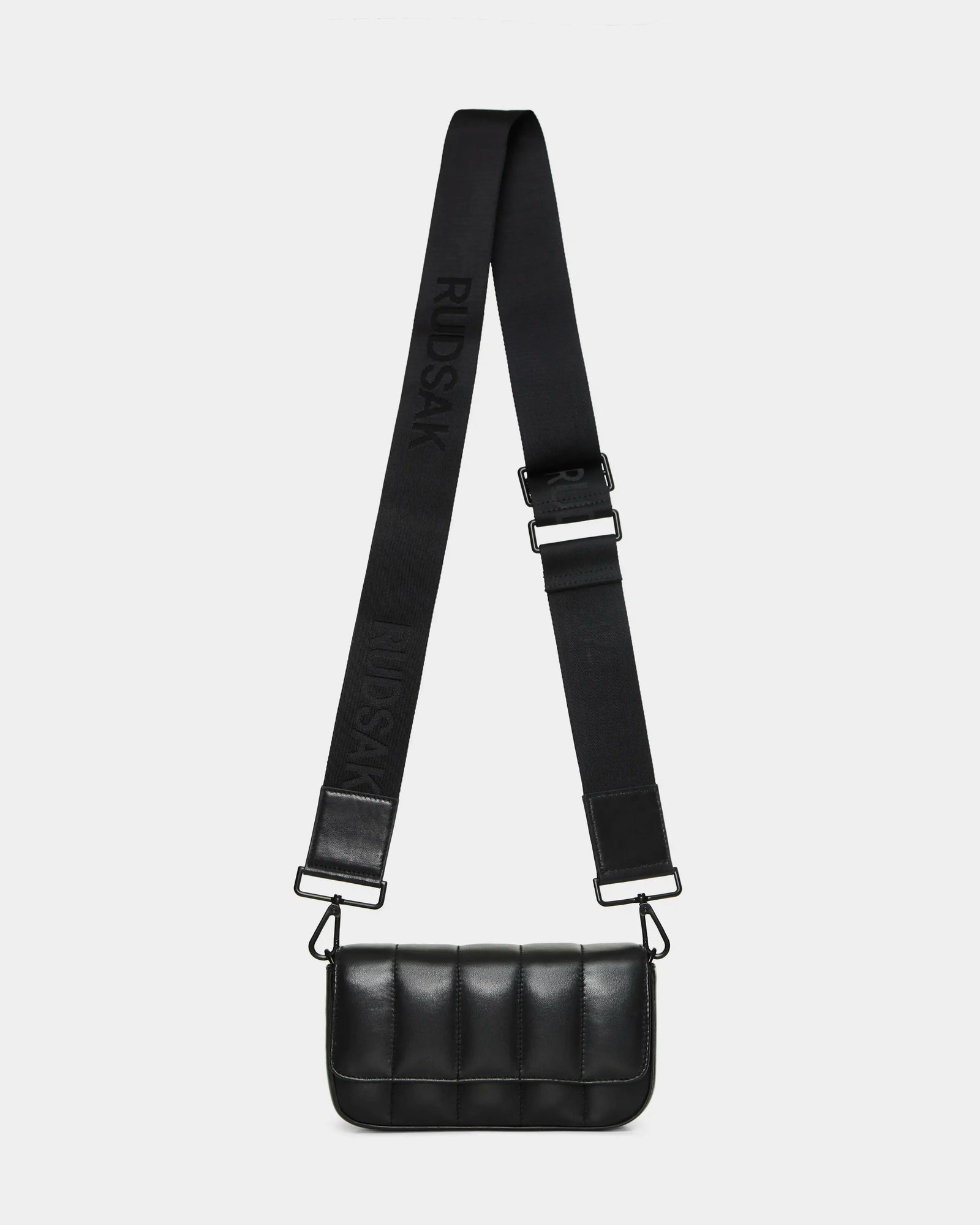 The CHARLI X - 8324009 Black by Rudsak is a small black quilted leather crossbody bag with a wide adjustable strap embossed with the brand name, sleek metal hardware, and a minimalist rectangular design.