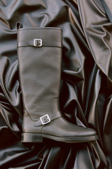 Gray leather knee-high boot with metal buckle straps, displayed on draped fabric background.