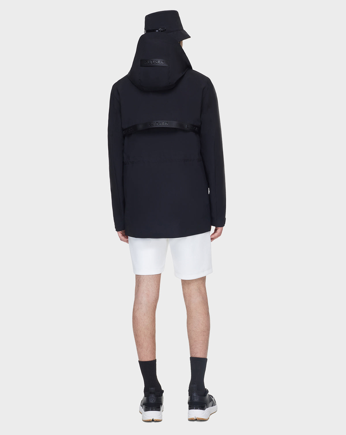 A person wearing the RUDSAK JACY - 6122000 BLACK trench, with a black bucket hat, white shorts, black socks, and black sneakers stands facing away against a plain white background.