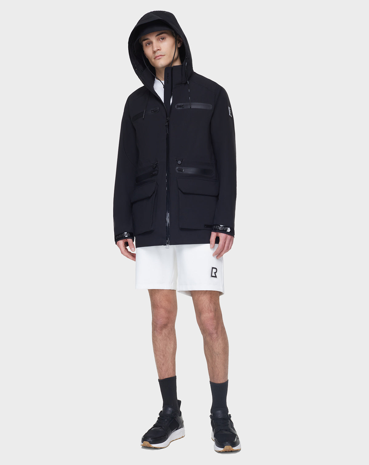 A person wearing the RUDSAK JACY - 6122000 BLACK rainwear jacket, white shirt, white R-logo shorts, black socks, and black sneakers stands against a plain light gray background.