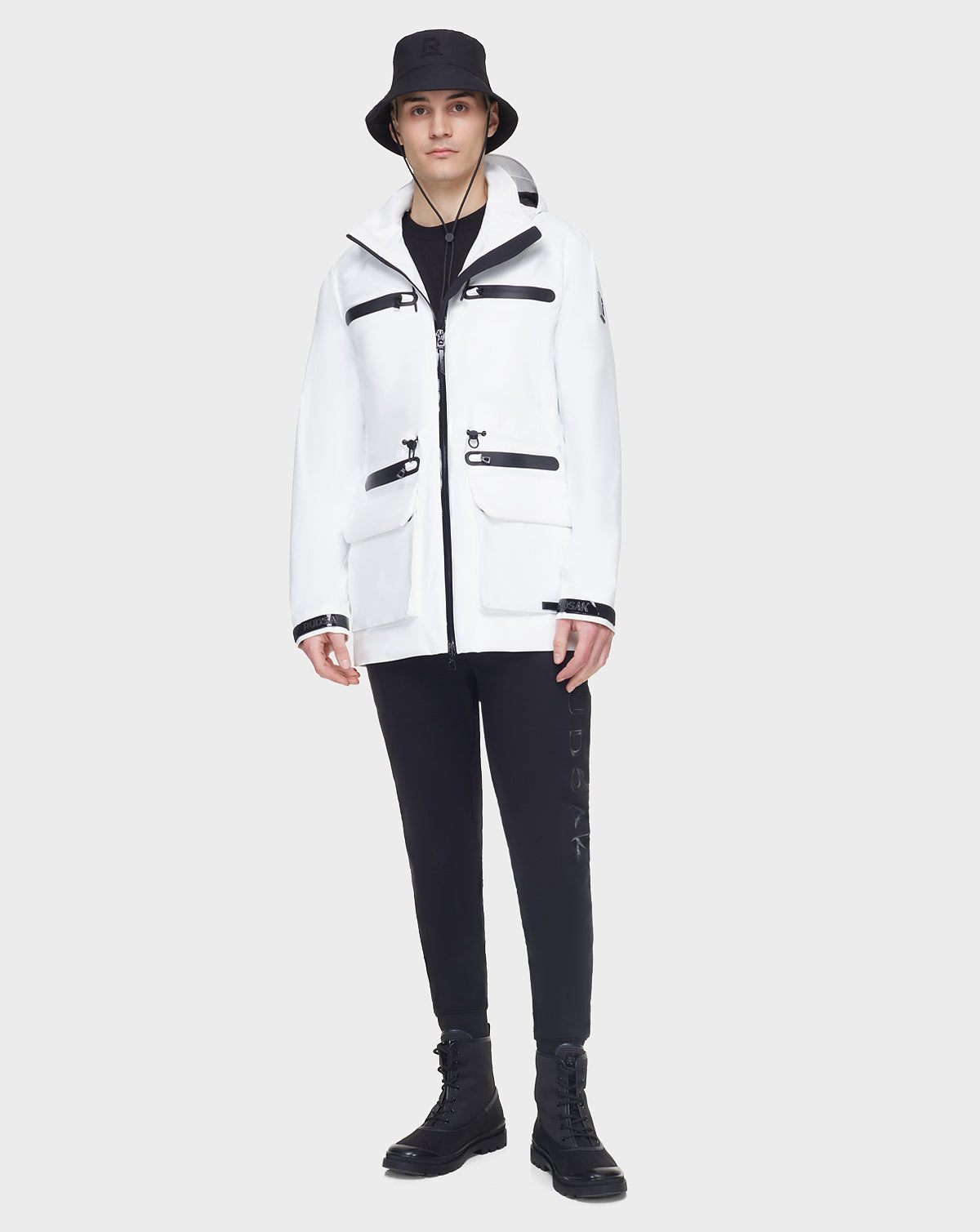A person stands against a light gray background, wearing the RUDSAK JACY - 6122000 WHITE mid-length raincoat with black zippers, styled with a black bucket hat, pants, and boots.