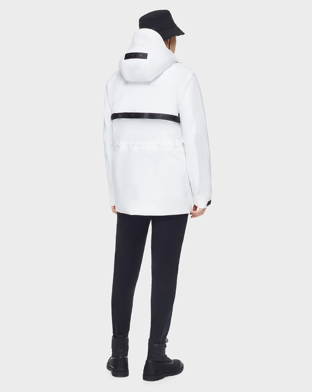 A person in the RUDSAK JACY - 6122000 WHITE water-resistant trench coat, paired with black pants, boots, and a bucket hat, faces away from the camera against a plain background.