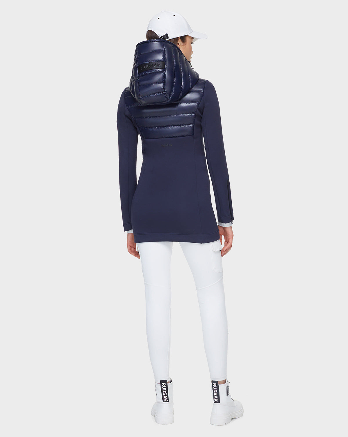 A woman faces away, wearing the Rudsak MARRY - CA EN L121003 DEEP BLUE puffer jacket with shiny hood, paired with white leggings, sneakers, and cap. The activewear-inspired look stands out against a plain light gray background.