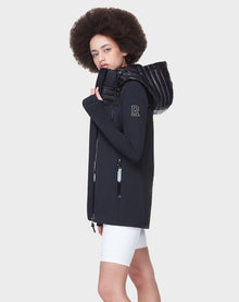 A person with a curly afro wears the Rudsak CAMIRA - 8122011 BLACK activewear jacket, featuring waterproof zippers and a padded collar, paired with white shorts, against a plain light gray background.